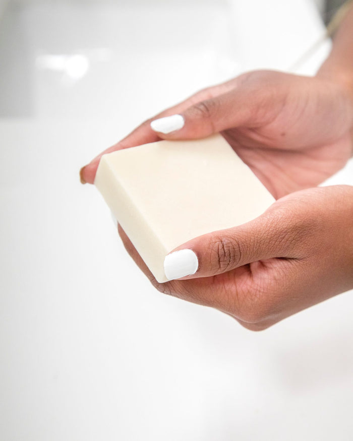 Two hands with white-painted nails hold a Lavender Sugar Scrub Bar by Spinster Sisters Co. over a sink. This off-white, rectangular bar is ideal for a plastic-free skincare routine, enhancing gentle exfoliation and hydration against the blurred background.