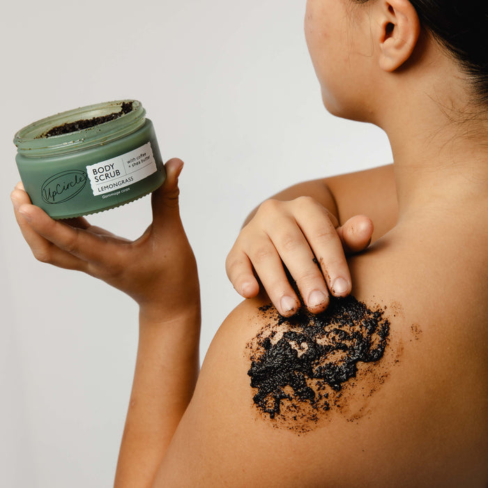 A person applies UpCircles Lemongrass Body Scrub with Coffee + Shea Butter to their shoulder, holding a round jar of the product against a plain white background.
