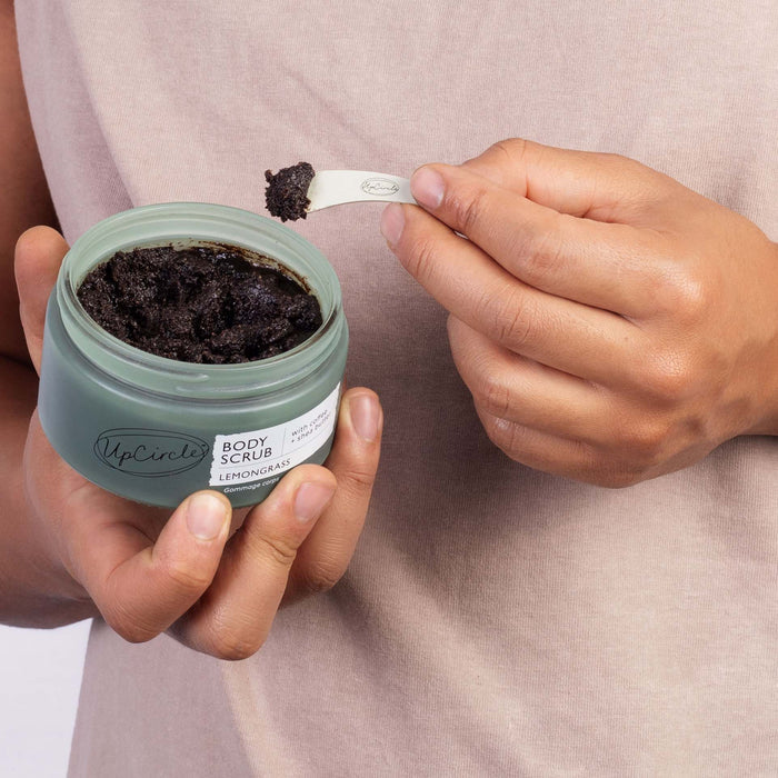 A person in a light beige top holds UpCircles Lemongrass Body Scrub with Coffee + Shea Butter in a green jar, using a small reusable spatula to scoop the sea salt-infused exfoliant.