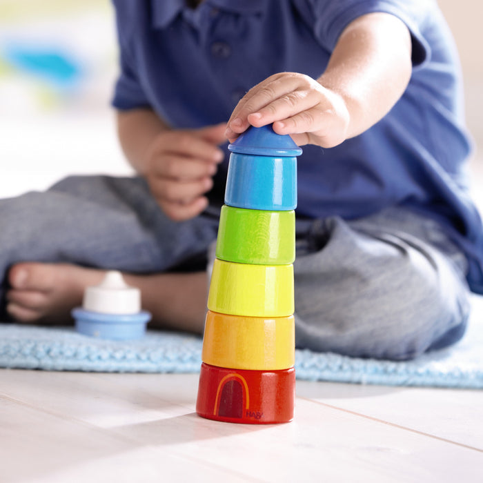 A child in a blue shirt sits on the floor, improving motor skills by stacking colorful cups made from sustainably sourced materials, starting with red and ending with blue. Next to a blue mat on the light floor is HABAs Lighthouse Wooden Rainbow Stacker.