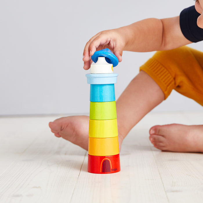 On a light wooden floor, a toddler in yellow pants and a dark shirt develops motor skills while assembling HABAs Lighthouse Wooden Rainbow Stacker, made of sustainably sourced materials. The toys colorful rings and blue top gleam as the child plays barefoot.