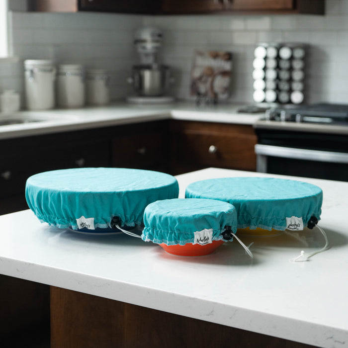 On a kitchen counter with white tiles and brown cabinets, three bowls in varying sizes are topped with the Linen Bowl Cover Bundle - Aqua by Marleys Monsters, creating a sustainable scene amid jars and a mixer.