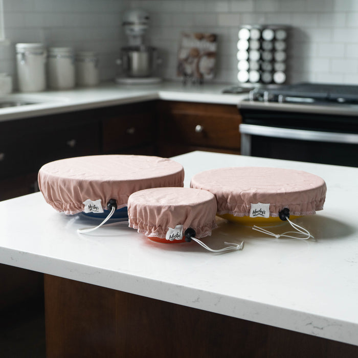 Three kitchen bowls in different sizes are covered with Marleys Monsters Linen Bowl Cover Bundle - Rose on a white countertop, with dark brown cabinets and appliances highlighting these eco-friendly, reusable solutions.