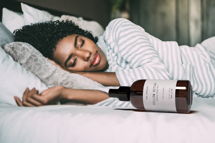 A person with curly hair sleeps peacefully on a bed with white sheets and a striped pillow. In the foreground, theres a bottle of Muse Apothecarys Linen, Pillow and Fabric Spray, suggesting its calming mist infused with natural ingredients is nearby.
