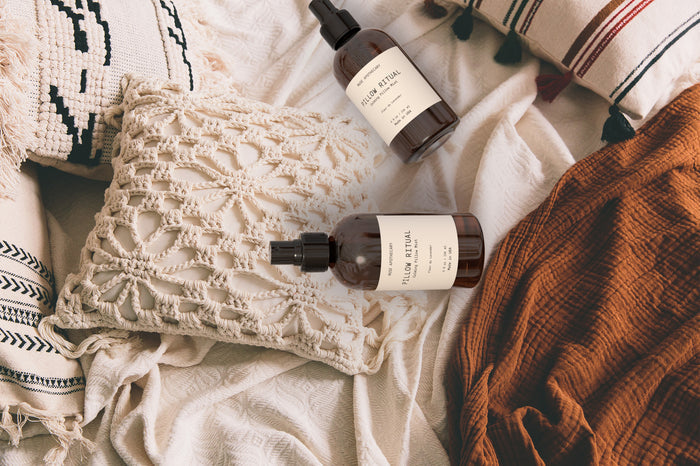 Two amber spray bottles labeled Linen, Pillow and Fabric Spray by Muse Apothecary rest on a boho-style bed with crocheted striped pillows and a textured orange blanket on an off-white duvet.