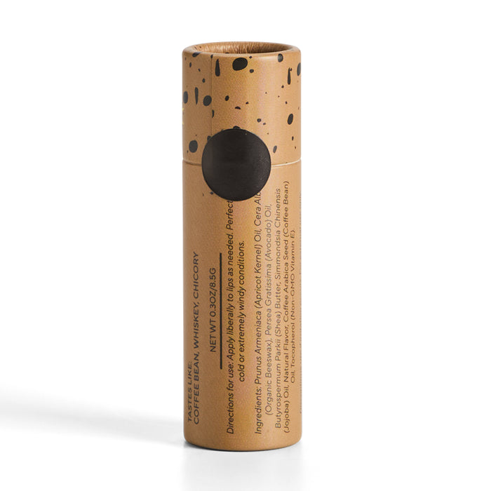 A brown cylindrical cardboard container with black ink splatters holds a lip balm from Soap Distillery. The package lists directions and ingredients, including apricot kernels, oils, and waxes. The label features coffee + whiskey notes with hints of mango coconut.