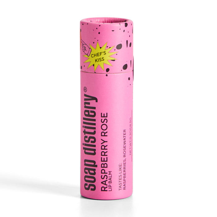 The Lip Balm by Soap Distillery comes in a pink cylindrical tube with splatter designs and a Chefs Kiss graphic. It boasts notes of raspberry, rosewater, and hints of coffee and whiskey sophistication.