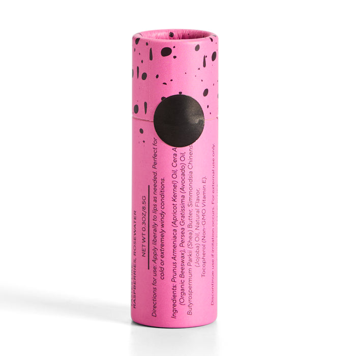A pink cylindrical Lip Balm container by Soap Distillery features black speckles and a round black label. The side displays directions, ingredients, and product details. Its matching pink cap with speckles perfectly complements the mango coconut flavor.