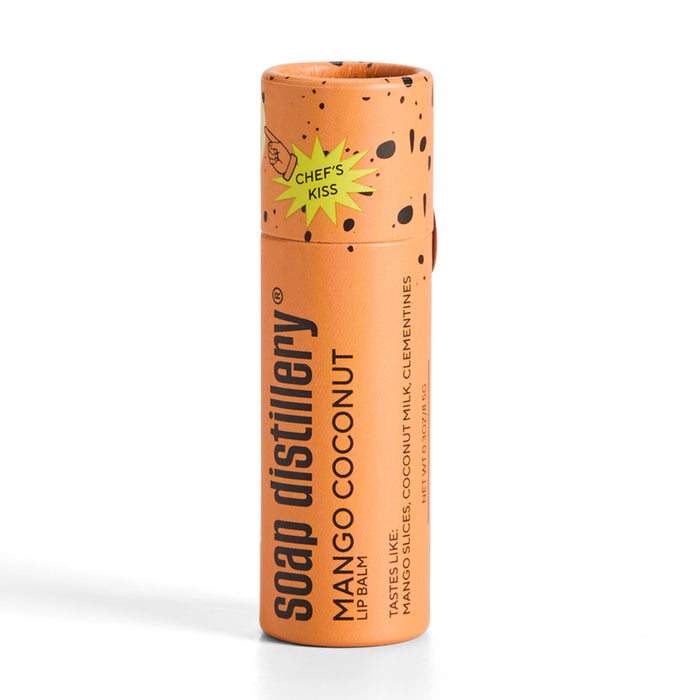 The vibrant orange cylindrical Lip Balm tube features the brand Soap Distillery and the scent Mango Coconut, adorned with a cheerful Chefs Kiss in a starburst. Experience luscious mango, creamy coconut milk, and clementines—ideal for a tropical escape on your lips!.