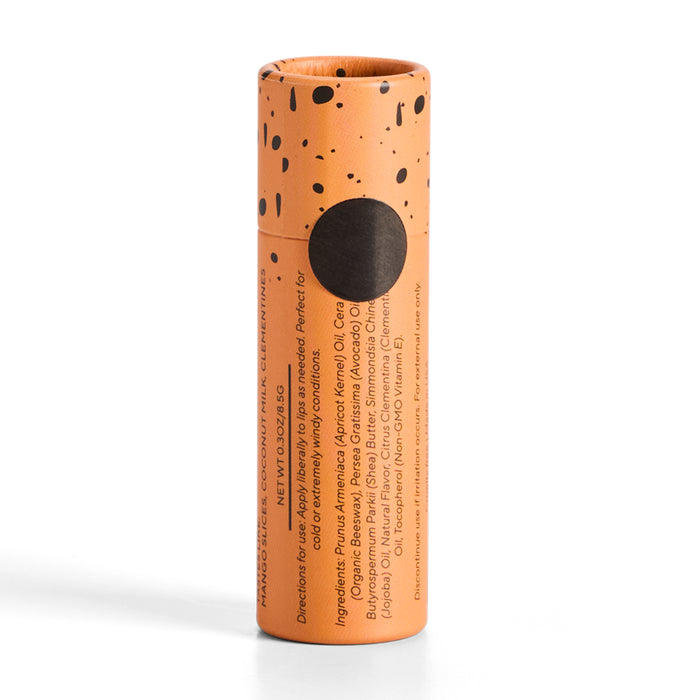 Orange cylindrical container with black speckles and a black circle, reminiscent of coffee + whiskey. Vertical text shows ingredients and directions. Cap is on. Product: Lip Balm by Soap Distillery.