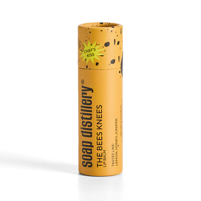 A cylindrical yellow Soap Distillery Lip Balm called The Bees Knees features lemon, honey, and juniper. It has natural ingredients, playful black speckles on the packaging, and a Chef’s Kiss graphic near the top.