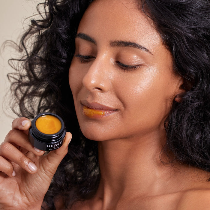 A curly-haired woman applies Henné Organics Lip Mask from a small black container to her lips. Made with organic ingredients, it nourishes and revitalizes her lips against a light, neutral backdrop.