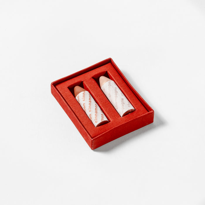 A red rectangular box holds two Axiology Balmie Duo crayons in branded paper, showcasing their vibrant vegan magic. The open box rests invitingly on a pristine white surface.