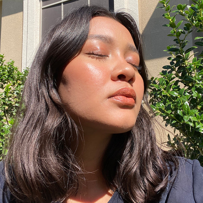 A person with long, dark hair stands outside with eyes closed, bathed in sunlight. Theyre wearing a dark top that complements the glow of Axiologys Balmie Duo. Green bushes and a building are visible in the background, highlighting their serene expression.