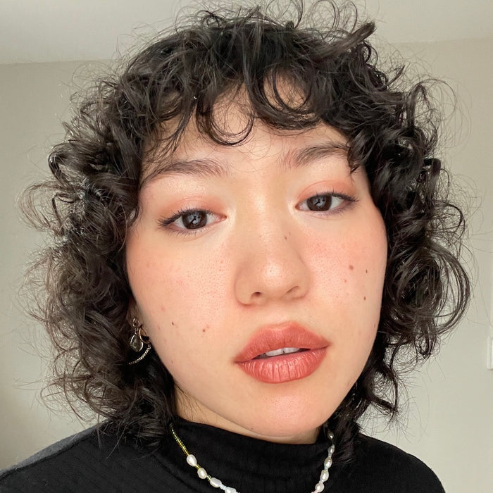 A person with curly hair and natural makeup looks at the camera expressionlessly. Wearing a black top, pearl necklace, and Axiologys Balmie Duo adds a touch of vegan charm against a plain indoor backdrop.