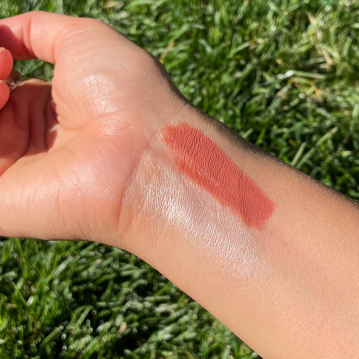A hand rests on grass showing two Axiology Balmie Duo swatches on the wrist: a shimmery white and a matte terracotta. These vegan multi-use crayons effortlessly elevate your beauty routine.