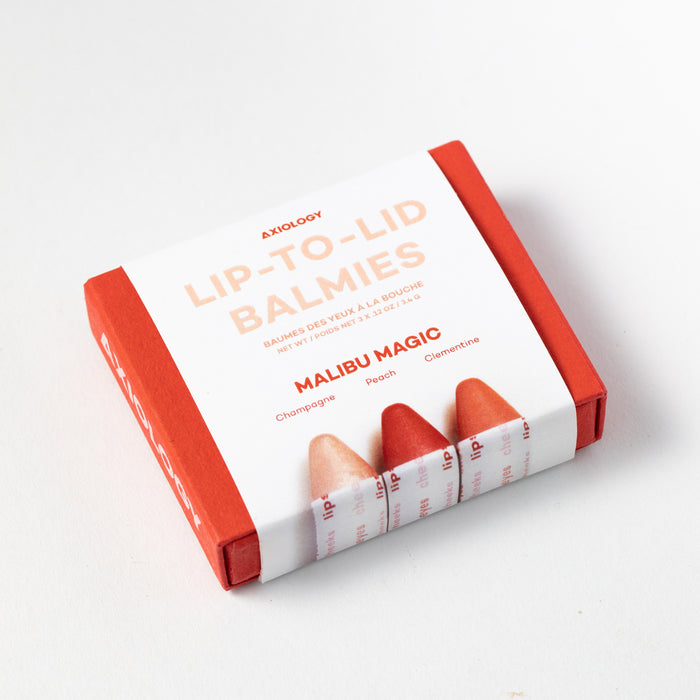 A square box labeled Balmie Trio by Axiology rests on a surface. The vegan red and white box features three multi-use crayon-like balms in Champagne, Peach, and Clementine, all under the enchanting name Malibu Magic.