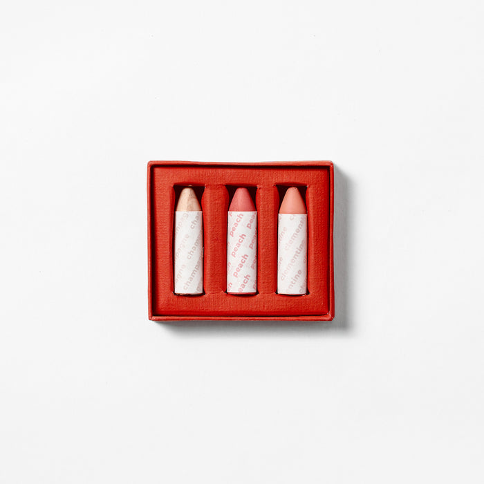 Axiologys Balmie Trio is presented in a red box and consists of three vegan, multi-use crayons in light beige and pink hues. They are vertically arranged, each wrapped in paper featuring red text on a plain white background.