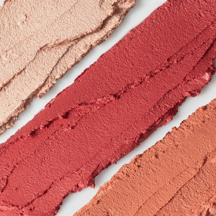 Three diagonal swatches on a white background display Balmie Trios creamy matte lipstick shades: light pink, deep red, and muted orange. Achieve this versatile look with Axiologys vegan Lip-to-Lid Balmies.