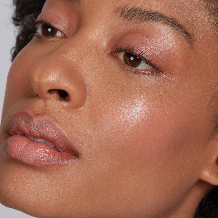 Close-up of a person with glowing skin enhanced by Axiologys vegan, natural Balmie Trio. Focus is on their smooth, radiant complexion using these multi-use crayons for subtle blush and neutral-toned lips. Lighting highlights the skins natural sheen.