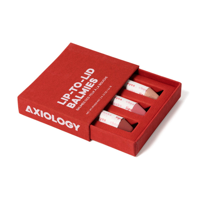 An open Axiology Lip-to-Lid Balmies Set - Cotton Candy Skies box reveals three versatile balm sticks in varying shades, partially exposed. Nestled on a white surface, the set comes in a recyclable case, embodying eco-friendly zero waste values.