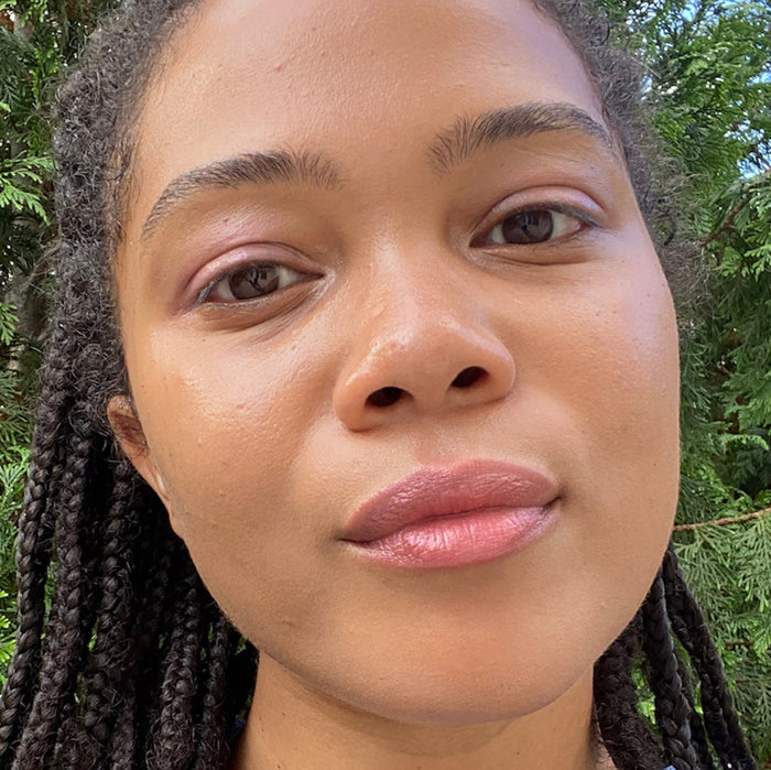Amid leafy green trees, a person with long braided hair and minimal makeup exudes natural beauty with Axiologys Lip-to-Lid Balmies Set in Cotton Candy Skies. Their neutral expression shines in bright light, highlighting features while embracing zero-waste practices.