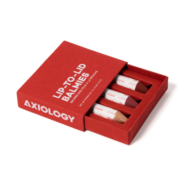 Open the red box to discover Axiologys Lip-to-Lid Balmies Set - Of the Earth, offering three vegan balmies ideal for a zero waste makeup routine. These versatile shades are crafted for eyes, cheeks, and lips, with white text on the box highlighting the product type and brand.
