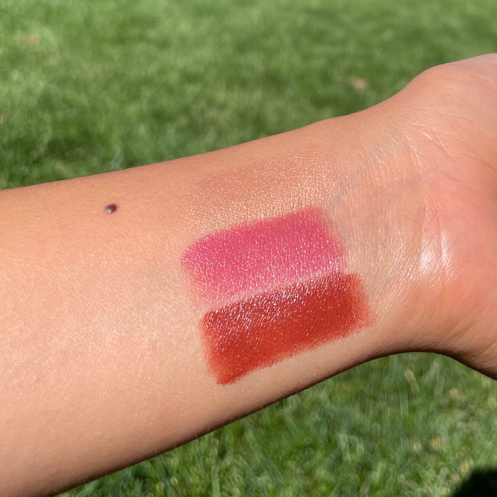 Two swatches from Axiologys Lip-to-Lid Balmies Set - Of the Earth, known for its zero waste makeup, are shown on an arm. The top shade is pink and the bottom is deep red, set against a backdrop of sunlit green grass.