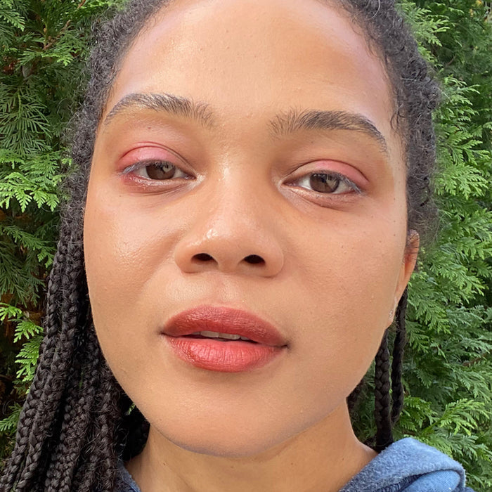 A person with braided hair and natural beauty stands outdoors surrounded by lush green foliage, their calm expression enhanced by Axiologys eco-friendly Lip-to-Lid Balmies Set - Of the Earth and a blue top, reflecting an elegant zero waste aesthetic.