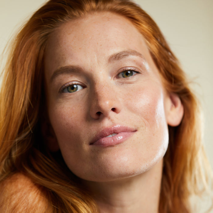 A person with long red hair smiles softly, displaying light skin and freckled cheeks. Their glossy lips shine from using Kari Grans Lip Whip Color Balm, against a blurred background that highlights the serene expression accentuated by its natural ingredients.