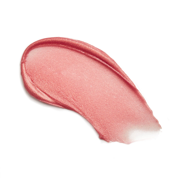 A smooth, shiny swipe of Kari Gran Lip Whip Color Balm in pink with a hint of shimmer on a white background reveals a creamy, slightly translucent texture crafted from natural ingredients.