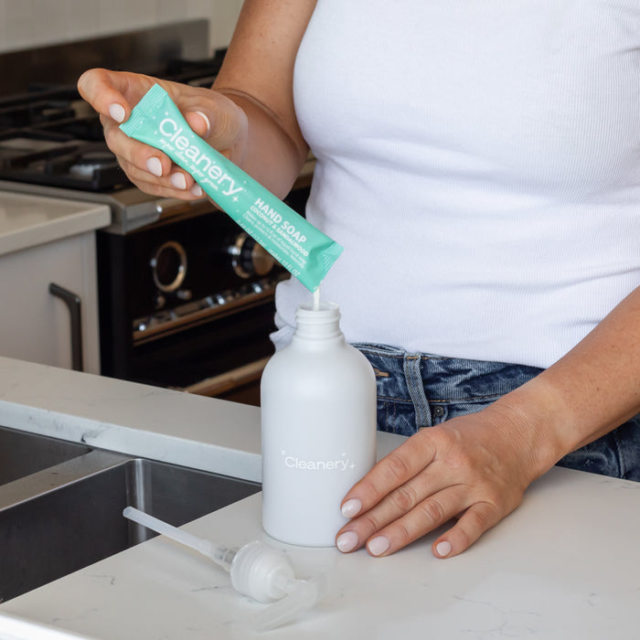 Wearing a white shirt, someone uses the Cleanery Liquid Hand Soap Starter Kit to dispense its green soap concentrate, perfect for sensitive skin, into a branded white bottle. Theyre in the kitchen near the sink and stove, with refills and a pump nearby on the counter.