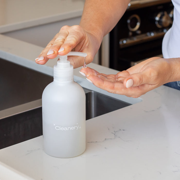 Someone with sensitive skin uses the Cleanery Liquid Hand Soap Starter Kit pump bottle to dispense soap onto their hand over the kitchen counter by the sink.