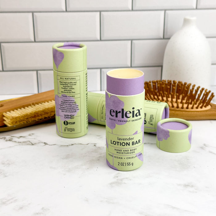 Two green and purple cylindrical lotion bars labeled Erleia Lotion Bar rest elegantly on a marble surface. Designed as hand and body moisturizers, they feature balm inside plastic-free packaging. A hairbrush and a white vase sit against a white subway tile wall in the background.