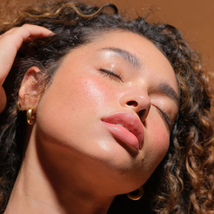 A woman with curly hair tilts her head back, eyes closed, showing radiant, dewy skin illuminated by RMS Beautys Champagne Rosé Luminizer. She touches her hair against a rich brown background.