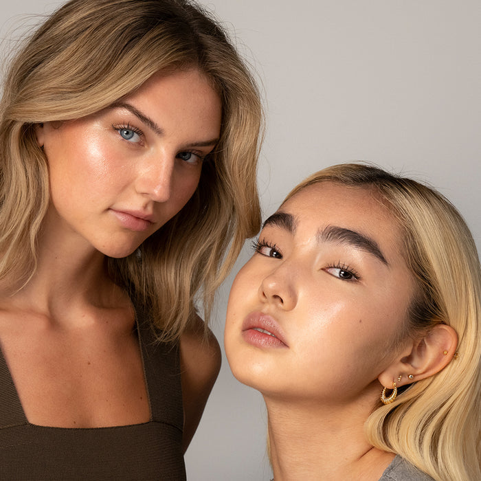 Two women with glowing skin pose together; one, in a brown top with wavy blonde hair and blue eyes, the other wearing gold hoops and gray top, achieves luminous skin using RMS Beauty’s Champagne Rosé Luminizer cream highlighter.