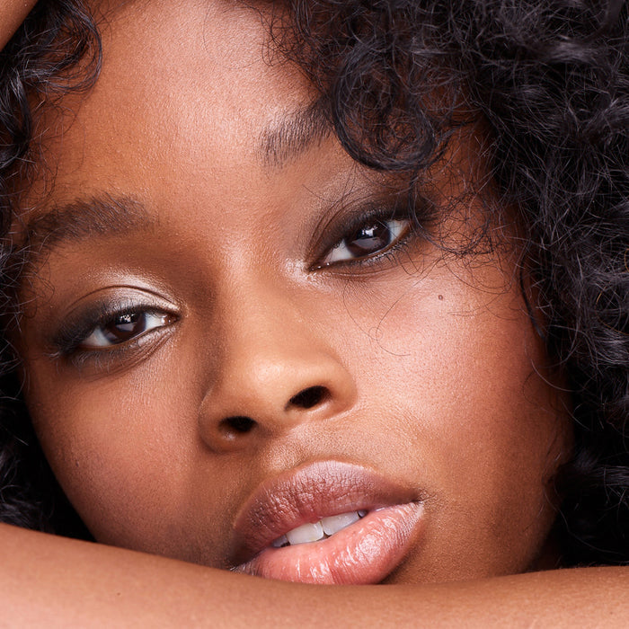 Close-up of a woman with curly hair and glowing skin, wearing natural makeup with subtle eye shadow, glossy lips, and a touch of RMS Beauty Champagne Rosé Luminizer for a dewy glow.
