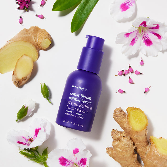 A purple bottle of Ursa Major Lunar Bloom Retinal Serum sits among pink and white flowers and ginger on a white background, ideal for enhancing skin texture and reducing fine lines.