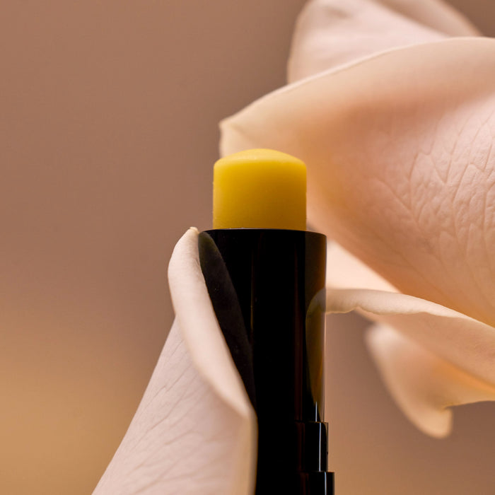 A close-up of the Luxury Lip Balm tube by Henné Organics, featuring a yellow balm partially surrounded by soft, pale pink rose petals. The blurred background enhances the contrast between the vibrant balm and delicate petals—ideal for natural skincare enthusiasts.