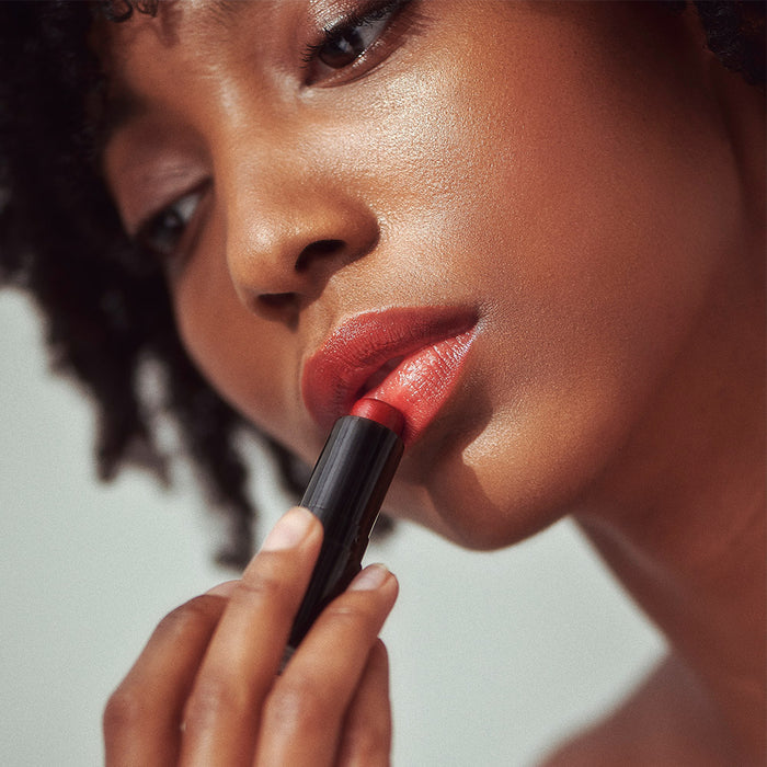 A close-up shows someone applying Henné Organics Luxury Lip Tint, focusing on lips and tint. Soft lighting highlights smooth skin and natural hair. The blurred background adds intimacy and elegance, showcasing the sophistication of this natural lip makeup.