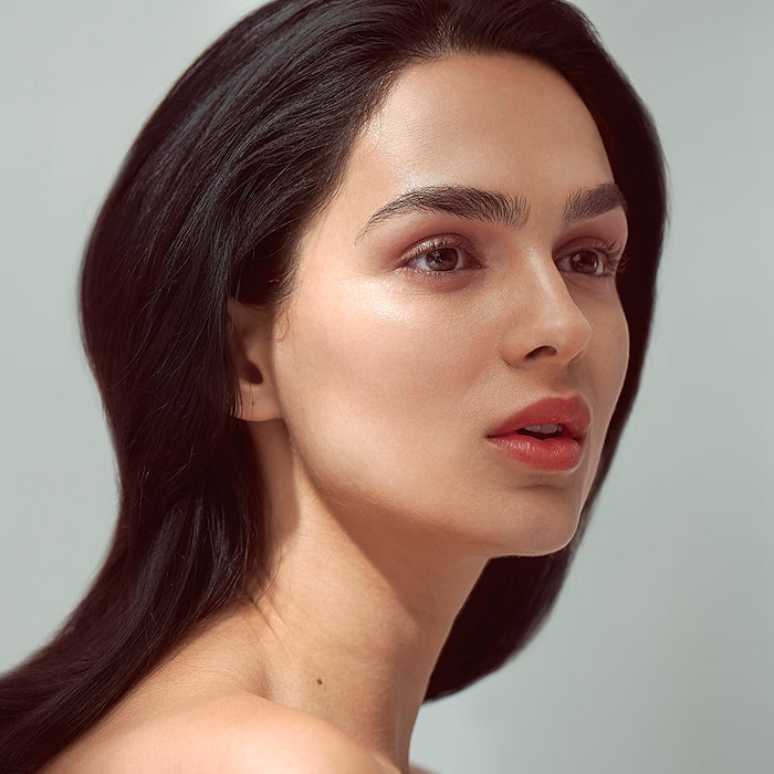 A woman with long black hair looks to the side, her smooth skin and elegantly tinted lips with Henné Organics Luxury Lip Tint. Her subtle makeup enhances defined eyebrows, set against soft light tones.