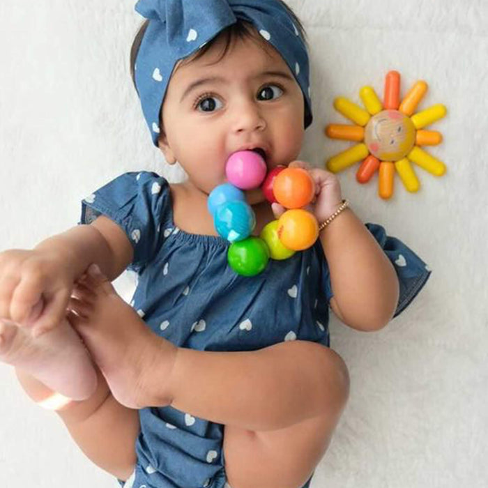A baby wearing a blue outfit with white hearts and a matching headband lies on a white surface, holding colorful balls, one in their mouth. Next to them is the non-toxic Magica Wooden Clutching Toy by HABA, adding playful charm to this delightful scene.