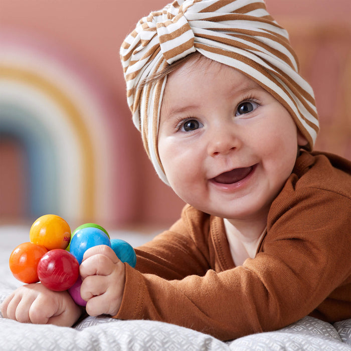 A smiling baby in a brown striped turban lies on a cushion, clutching the colorful HABA Magica Wooden Clutching Toy. A rainbow decoration blurs in the background as the baby sports a brown outfit.