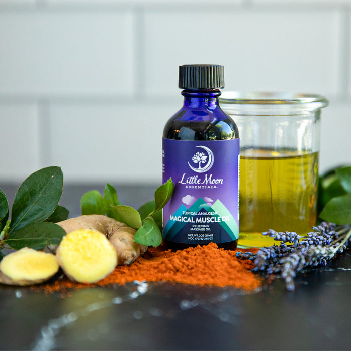 The bottle of Little Moon Essentials Magical Muscle Oil™ is surrounded by fresh ginger, green leaves, lavender, and spices—natural pain relievers. Nearby sits a small jar of oil on a dark surface against light tiles.