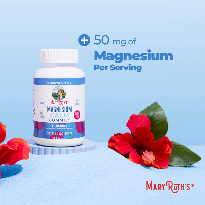 A bottle of MaryRuth's Magnesium Calm Gummies rests on blue sand with hibiscus flowers, showcasing "50 mg of Magnesium Per Serving" above. Ideal for promoting bone health and muscle relaxation, the scene is framed by a calming gradient blue backdrop.