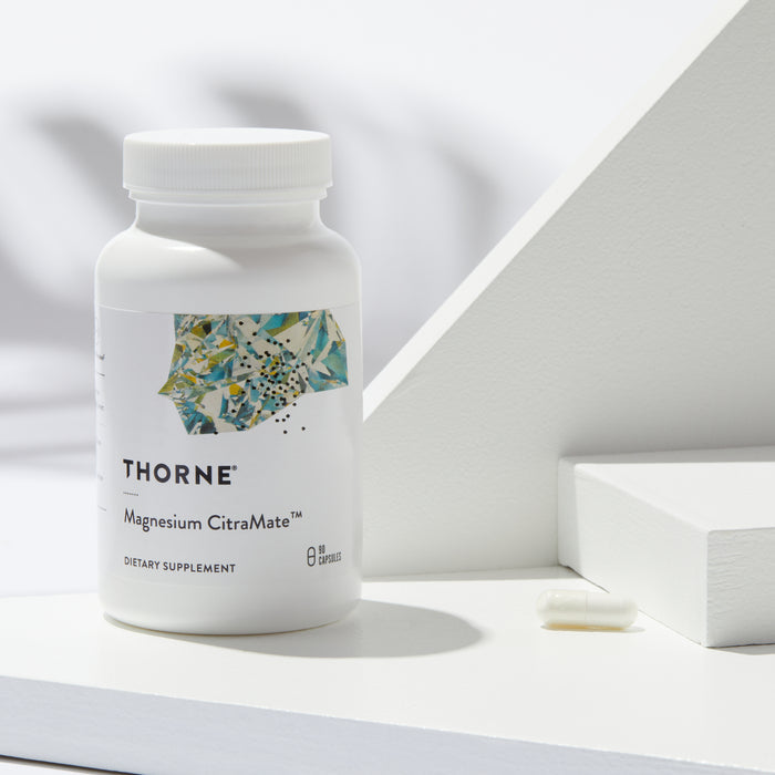 A bottle of Thornes Magnesium CitraMate dietary supplement sits on a white surface with geometric accents, emphasizing its muscle support benefits. Beside it, a capsule rests while the label displays an abstract design in soothing green and blue tones.