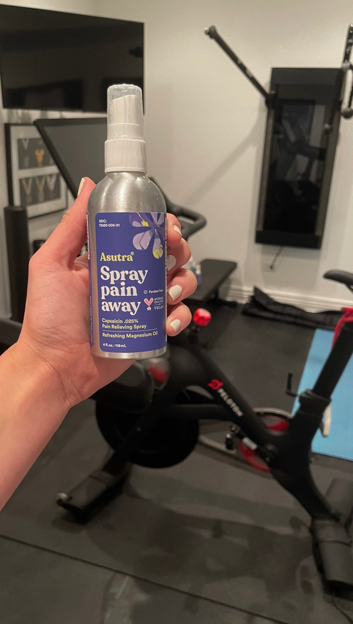 In the foreground, someone holds a bottle of Asutra Magnesium Oil Spray, known for relieving muscle cramps. The background features an exercise bike, weights, and gym equipment in a well-equipped home gym setting.