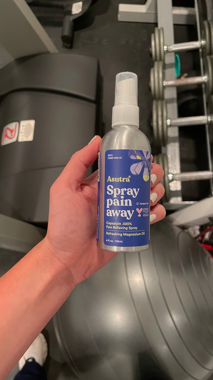 A hand holds a bottle of Asutras Magnesium Oil Spray, perfect for relieving muscle cramps. In the background, gym equipment like a treadmill, exercise ball, and dumbbells emphasize its ideal use in fitness routines.