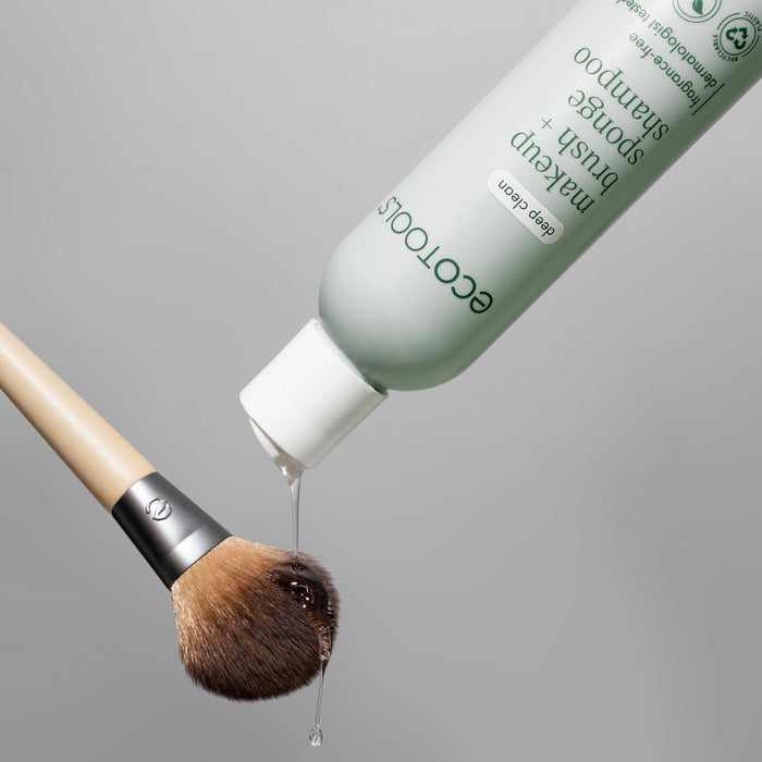 An EcoTools Makeup Brush & Sponge Shampoo bottle, celebrated for its paraben-free formula, is tilted to pour liquid onto a fluffy brush against a plain gray background.