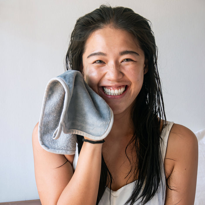 A smiling person with wet hair holds Tree To Tubs gray, hypoallergenic Makeup Remover Towel to their cheek. Wearing a white sleeveless top and sitting indoors against a light background, they enjoy the towels gentle cleanse.